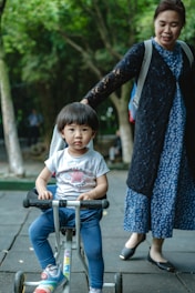 A young child sits on a small tricycle while an adult stands nearby. The adult is wearing a blue dress with a black cardigan. The background features green trees, suggesting an outdoor setting.
