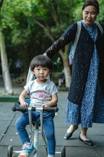 A young child sits on a small tricycle while an adult stands nearby. The adult is wearing a blue dress with a black cardigan. The background features green trees, suggesting an outdoor setting.