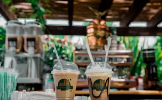 Two iced coffee drinks in plastic cups with straws are placed on a wooden counter, surrounded by a lush, green backdrop. In the background, there is a coffee machine and blurred greenery, creating a warm, inviting atmosphere.