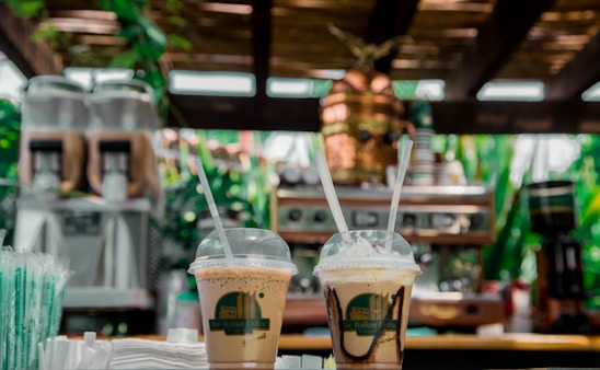 Two iced coffee drinks in plastic cups with straws are placed on a wooden counter, surrounded by a lush, green backdrop. In the background, there is a coffee machine and blurred greenery, creating a warm, inviting atmosphere.