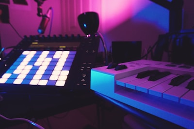 A music production setup featuring an electronic music pad controller with illuminated buttons and a MIDI keyboard. The setting is dimly lit with vibrant purple and pink lighting, creating a moody and creative atmosphere. Various equipment and cables are visible in the background.