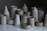 A collection of ceramic vessels, including cups, jugs, and pots, arranged on a reflective surface. The ceramics have a matte finish and are predominantly gray with subtle speckles.
