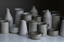 A collection of ceramic vessels, including cups, jugs, and pots, arranged on a reflective surface. The ceramics have a matte finish and are predominantly gray with subtle speckles.