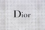 The image features the word 'Dior' prominently displayed in bold black letters against a patterned background. The background is composed of intricate, repeating geometric designs in navy blue and white tones, giving an elegant and sophisticated appearance.