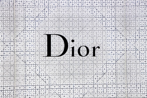 The image features the word 'Dior' prominently displayed in bold black letters against a patterned background. The background is composed of intricate, repeating geometric designs in navy blue and white tones, giving an elegant and sophisticated appearance.