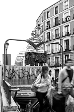 A black and white image depicting the entrance to the Ant&oacute;n Mart&iacute;n metro station in an urban setting. Blurred figures of people are descending the stairs, indicating movement and the hustle of city life. A statue of several figures stands prominently above the entrance, and a graffiti-marked fence is visible in the background. The buildings lining the street exhibit classic architecture with multiple balconies.
