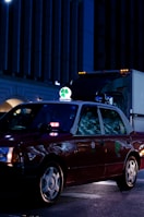 A dark red taxi is parked on a city street at night, illuminated by streetlights and building lights. The taxi has a bright green and white sign on top featuring a four-leaf clover. Reflections from the surroundings are visible on the car's windows.