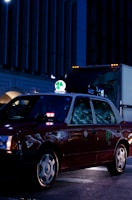 A dark red taxi is parked on a city street at night, illuminated by streetlights and building lights. The taxi has a bright green and white sign on top featuring a four-leaf clover. Reflections from the surroundings are visible on the car's windows.
