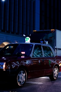 A dark red taxi is parked on a city street at night, illuminated by streetlights and building lights. The taxi has a bright green and white sign on top featuring a four-leaf clover. Reflections from the surroundings are visible on the car's windows.