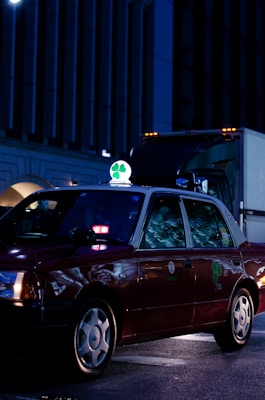 A dark red taxi is parked on a city street at night, illuminated by streetlights and building lights. The taxi has a bright green and white sign on top featuring a four-leaf clover. Reflections from the surroundings are visible on the car's windows.