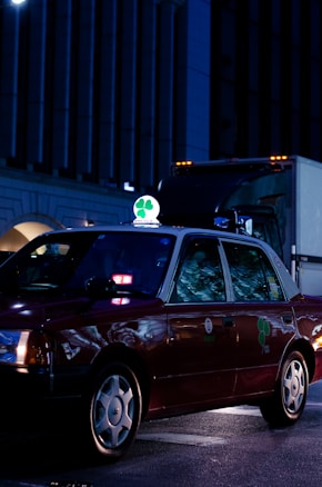 A dark red taxi is parked on a city street at night, illuminated by streetlights and building lights. The taxi has a bright green and white sign on top featuring a four-leaf clover. Reflections from the surroundings are visible on the car's windows.