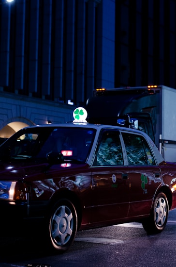 A dark red taxi is parked on a city street at night, illuminated by streetlights and building lights. The taxi has a bright green and white sign on top featuring a four-leaf clover. Reflections from the surroundings are visible on the car's windows.