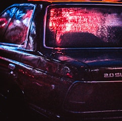A dark car with wet surfaces reflecting red and blue neon lights, creating an atmospheric and moody scene. The focus is on the rear of the vehicle, highlighting the branding and textures.