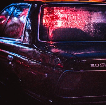 A dark car with wet surfaces reflecting red and blue neon lights, creating an atmospheric and moody scene. The focus is on the rear of the vehicle, highlighting the branding and textures.
