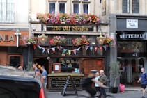 A traditional pub with a wooden facade adorned with colorful hanging flower baskets and vintage-style lanterns. A row of vibrant bunting decorates the entrance. The surrounding buildings display signage, including one with a 'To Let' sign. Several people are walking past, contributing to a lively street ambiance.