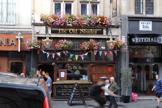 A traditional pub with a wooden facade adorned with colorful hanging flower baskets and vintage-style lanterns. A row of vibrant bunting decorates the entrance. The surrounding buildings display signage, including one with a 'To Let' sign. Several people are walking past, contributing to a lively street ambiance.