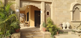 The image features an ornate entrance with intricate stone carvings and lattice work. Several potted plants and green foliage add natural elements to the setting. There are wooden chairs arranged in the porch area, providing a traditional and welcoming vibe. The architecture exhibits detailed patterns and a warm, earthy color palette.