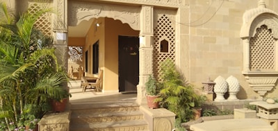 The image features an ornate entrance with intricate stone carvings and lattice work. Several potted plants and green foliage add natural elements to the setting. There are wooden chairs arranged in the porch area, providing a traditional and welcoming vibe. The architecture exhibits detailed patterns and a warm, earthy color palette.