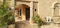 The image features an ornate entrance with intricate stone carvings and lattice work. Several potted plants and green foliage add natural elements to the setting. There are wooden chairs arranged in the porch area, providing a traditional and welcoming vibe. The architecture exhibits detailed patterns and a warm, earthy color palette.