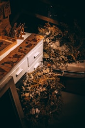 A dimly lit workspace features a white wooden cabinet adorned with vintage-style paper designs. On the cabinet top are various decorative dried plants and flowers, including some laid out in a wreath. The overall scene suggests a rustic, artistic setting with an emphasis on natural elements.