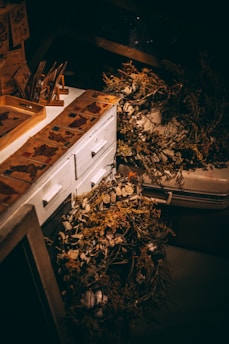 A dimly lit workspace features a white wooden cabinet adorned with vintage-style paper designs. On the cabinet top are various decorative dried plants and flowers, including some laid out in a wreath. The overall scene suggests a rustic, artistic setting with an emphasis on natural elements.