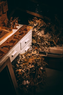 A dimly lit workspace features a white wooden cabinet adorned with vintage-style paper designs. On the cabinet top are various decorative dried plants and flowers, including some laid out in a wreath. The overall scene suggests a rustic, artistic setting with an emphasis on natural elements.