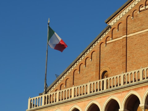 An Italian flag is waving atop a historic building with a brick facade. The building features arched windows and an ornamental balustrade. The sky is clear, providing a sharp contrast to the warm color of the bricks.
