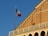 An Italian flag is waving atop a historic building with a brick facade. The building features arched windows and an ornamental balustrade. The sky is clear, providing a sharp contrast to the warm color of the bricks.