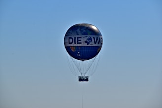 A hot air balloon with a design resembling a globe, floating in a clear blue sky. The balloon features the text 'DIE WELT' printed across its surface. The basket is visible beneath the balloon, tethered by numerous cables.
