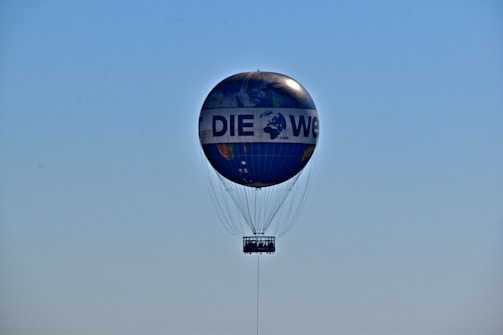 A hot air balloon with a design resembling a globe, floating in a clear blue sky. The balloon features the text 'DIE WELT' printed across its surface. The basket is visible beneath the balloon, tethered by numerous cables.