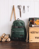 A green canvas backpack with leather straps is positioned against a white wooden wall. Next to it, a wooden crate labeled with various text, an amber glass bottle, and pieces of firewood are arranged. Above the backpack, there are vintage tools hanging on the wall, including scissors, pliers, a ruler, and a hook. The scene has a rustic, vintage aesthetic.