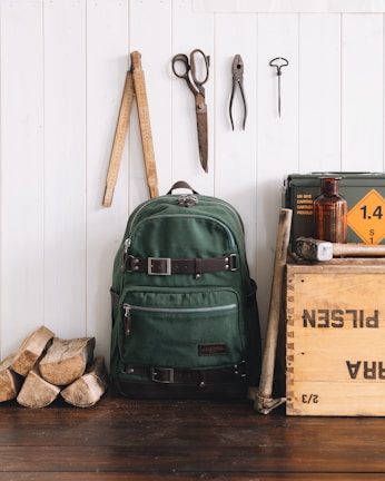 A green canvas backpack with leather straps is positioned against a white wooden wall. Next to it, a wooden crate labeled with various text, an amber glass bottle, and pieces of firewood are arranged. Above the backpack, there are vintage tools hanging on the wall, including scissors, pliers, a ruler, and a hook. The scene has a rustic, vintage aesthetic.
