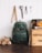 A green canvas backpack with leather straps is positioned against a white wooden wall. Next to it, a wooden crate labeled with various text, an amber glass bottle, and pieces of firewood are arranged. Above the backpack, there are vintage tools hanging on the wall, including scissors, pliers, a ruler, and a hook. The scene has a rustic, vintage aesthetic.