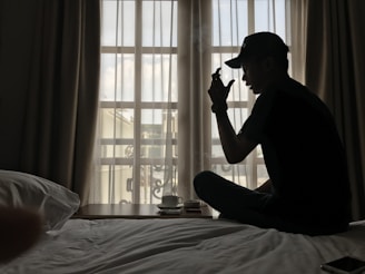 A person wearing a cap is sitting cross-legged on a bed next to a bedside table with a coffee cup. The room is softly lit by natural light coming through a window with sheer curtains.