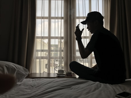 A person wearing a cap is sitting cross-legged on a bed next to a bedside table with a coffee cup. The room is softly lit by natural light coming through a window with sheer curtains.