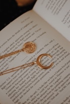 An open book displays text while serving as a background for jewelry. A gold necklace with a round medallion and a crescent moon design is elegantly placed across the pages, adding a touch of charm to the literary scene.