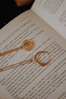 An open book displays text while serving as a background for jewelry. A gold necklace with a round medallion and a crescent moon design is elegantly placed across the pages, adding a touch of charm to the literary scene.