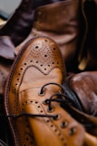 A close-up of a polished brown leather brogue shoe with intricate perforation details and laces, surrounded by other leather footwear in a slightly blurred background.