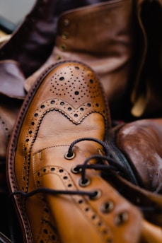 A close-up of a polished brown leather brogue shoe with intricate perforation details and laces, surrounded by other leather footwear in a slightly blurred background.