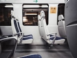 A row of empty seats inside a train or subway car, with blue and white upholstery. The interior is clean and modern, featuring metal armrests and windows reflecting the outside scene. A sign in the corner suggests reserved seating.