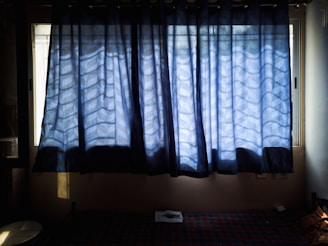 A pair of blue curtains with a wave pattern partially covers two tall windows, allowing some light to filter through. Below the curtains is a checkered bedspread, and there is a small table with a few items on it.