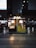 A small cafe setup inside a train station, with illuminated signage displaying '@Cafe'. The cafe has a variety of snacks and beverages displayed in glass cases. The surrounding area is bustling with people and is marked by directional signs and platform information.