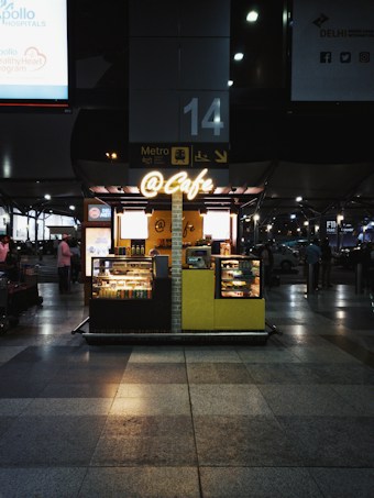 A small cafe setup inside a train station, with illuminated signage displaying '@Cafe'. The cafe has a variety of snacks and beverages displayed in glass cases. The surrounding area is bustling with people and is marked by directional signs and platform information.