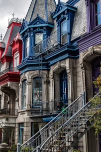A row of colorful townhouses features ornate architectural details with prominent use of rich colors. The houses are painted in red, blue, and purple, each with intricate trims and iron railings. The steep steps leading up to the doors add to the urban charm.
