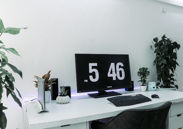A minimalistic workspace setup with a large digital clock displayed on a computer monitor. The desk is white and organized with a keyboard and mouse in front of the monitor. Various plants provide a touch of greenery. A small camera and other gadgets are also present on the desk.