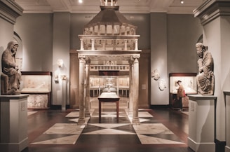A museum gallery displaying classical sculptures and artifacts. The room features stone statutes of seated figures on pedestals, a grand architectural structure with columns in the center, and various artifacts in glass cases along the walls. Soft lighting enhances the historical ambiance.