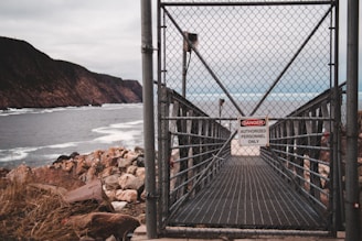 A chain-link fence gate with a sign reading 'Danger: Authorized Personnel Only' blocks access to a narrow walkway. The walkway appears to lead towards a rocky coastline with cliffs in the background. The sea is visible, with waves crashing against the rocks, and the sky is overcast.
