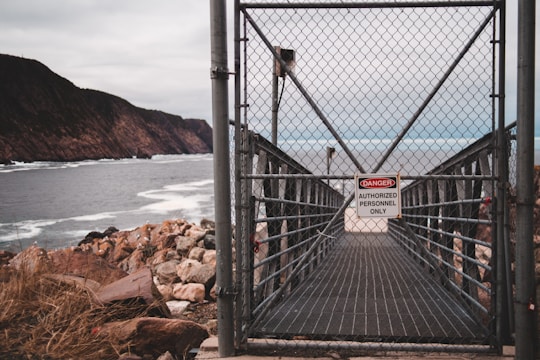 A chain-link fence gate with a sign reading 'Danger: Authorized Personnel Only' blocks access to a narrow walkway. The walkway appears to lead towards a rocky coastline with cliffs in the background. The sea is visible, with waves crashing against the rocks, and the sky is overcast.