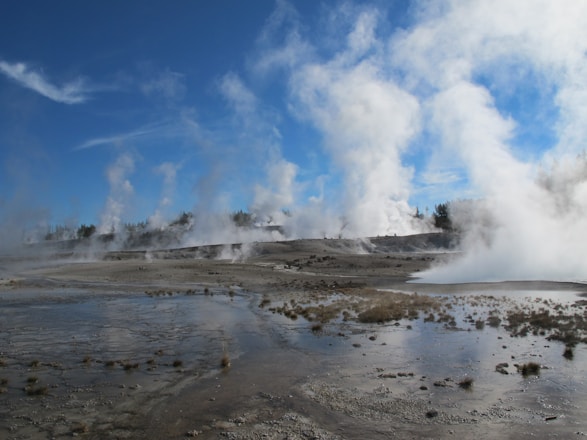 A geothermal landscape with numerous steam vents and hot springs emits plumes of vapor into a clear blue sky. The barren ground is intermixed with small pools of water, creating a dramatic and rugged natural scene.