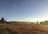 A vast open prairie landscape with golden brown grasses under a clear blue sky. In the distance, mountains are visible with some mist hanging above them. On the left, a small wooden cabin is nestled among a few tall pine trees. The image has a serene and peaceful quality.